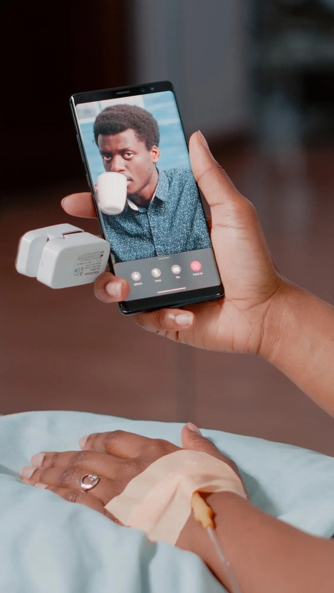 Молодая афроамериканская женщина, сидя в больничной палате, по видеосвязи со смартфона разговаривает с мужчиной. Пациентка с подключённой капельницей общается со взрослым мужчиной на онлайн-видеоконференции.